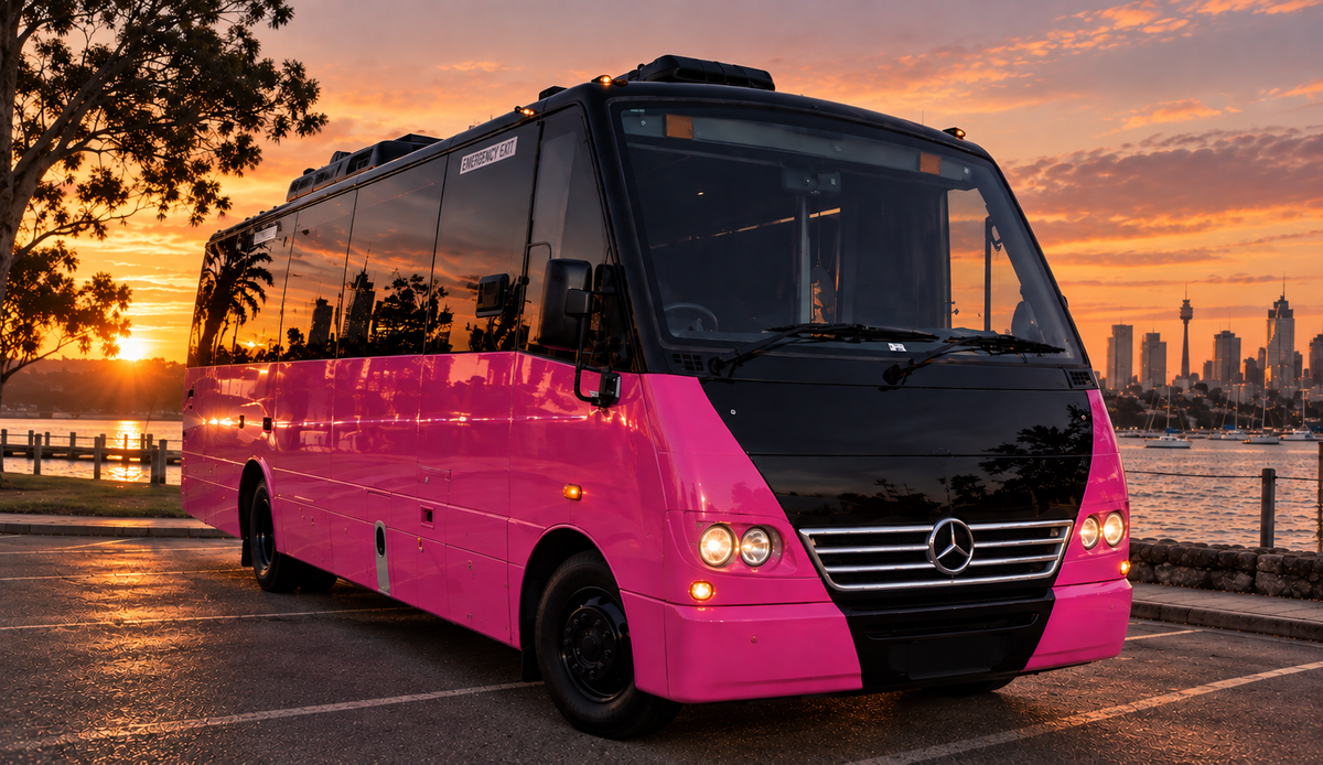 A pink and black Mercedes-Benz coach bus parked outdoors near a waterfront at sunset, with a city skyline and water visible in the background.