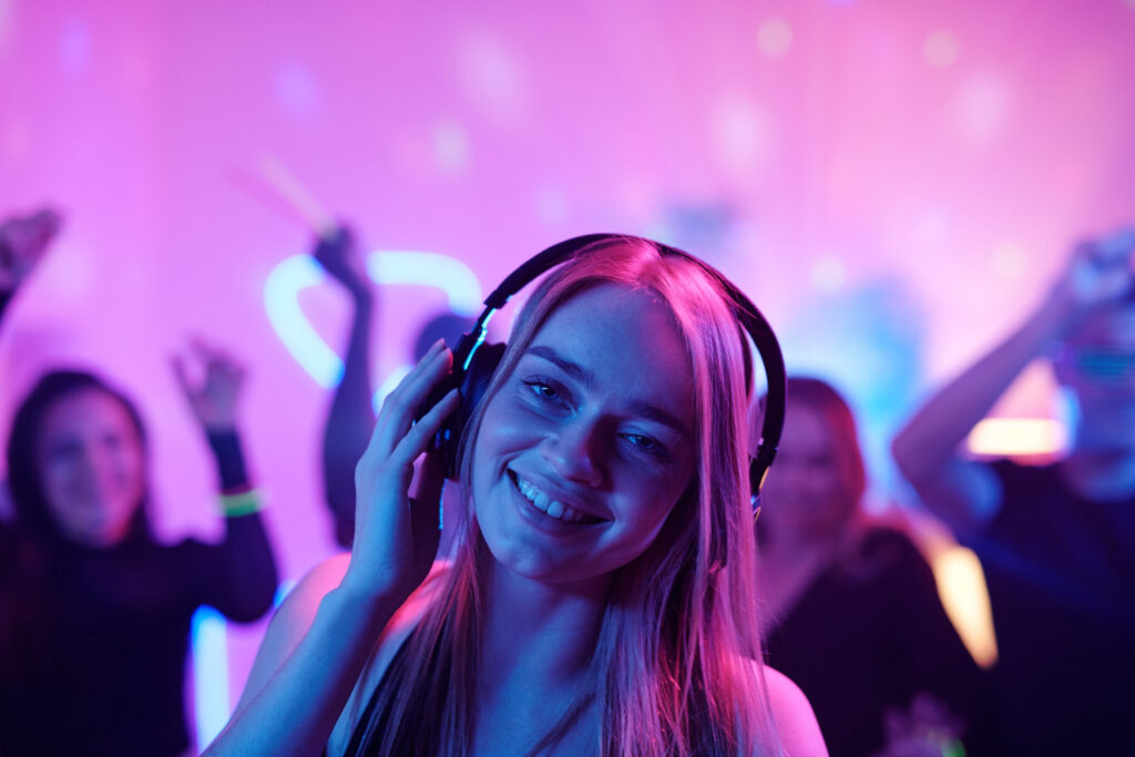 young cheerful woman with long blond hair snapping fingers while enjoying music headphones front dancing crowd party